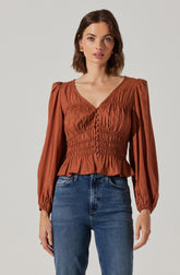 A woman is wearing a long-sleeved, rust-colored blouse with puffed shoulders and elasticized cuffs. The blouse features a V-neckline, a row of buttons down the front, and gathered detailing at the waist, creating a slightly peplum effect. She pairs the blouse with high-waisted, medium-wash blue jeans.